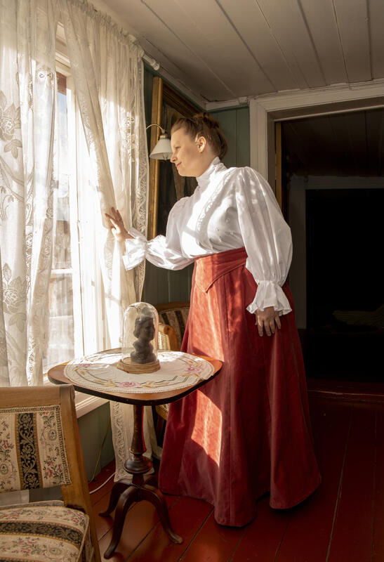 Ung kvinne iført hvit bluse og fotsidt, rødt skjørt med høyt liv står og kikker ut av et vindu med blonegardiner.