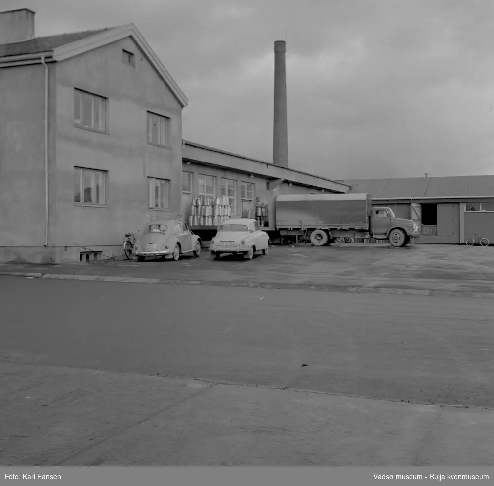A/L Aust-Finnmark Meieri, 1962 i Vadsø. Bildet er tatt mot nordøst. En lastebil er rygget inn til bygningen for av/pålasting og to personbiler står parkert på sydsiden av bygningen.
Arbeidet med å få meieri til Vadsø ble påbegynt før krigen.  Ønsket var å få et meieri som kunne dekke Nord-Varanger-Tanadistriktet.  Andelslaget A/L Aust-Finnmark Meieri ble stiftet 28.juni 1947 i Nyborg.  
Aust- Finnmark Meieri ble oppført i Tollbugata i 1949; arkitekt Monrad Heggen m hovedentreprenør Brdr. Aronsen.
Oppstart skulle finne sted ca 1. oktober 1949, og i mai ble det ansatt en meieriarbeider; Richard Korbi fra Vadsø.
Kilde: "A.L Aust-Finnmark Meieri 25 års jubileum 1949-1974; styreformann Ivar Johnsen, Tana mai 1974"