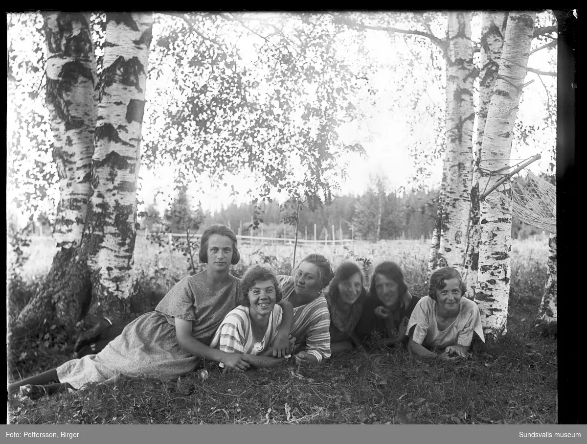 Sex unga kvinnor ligger i gräset på gården Ystibacken i Mjösund, Njurunda. På bild två sitter de och fikar i trädgården.