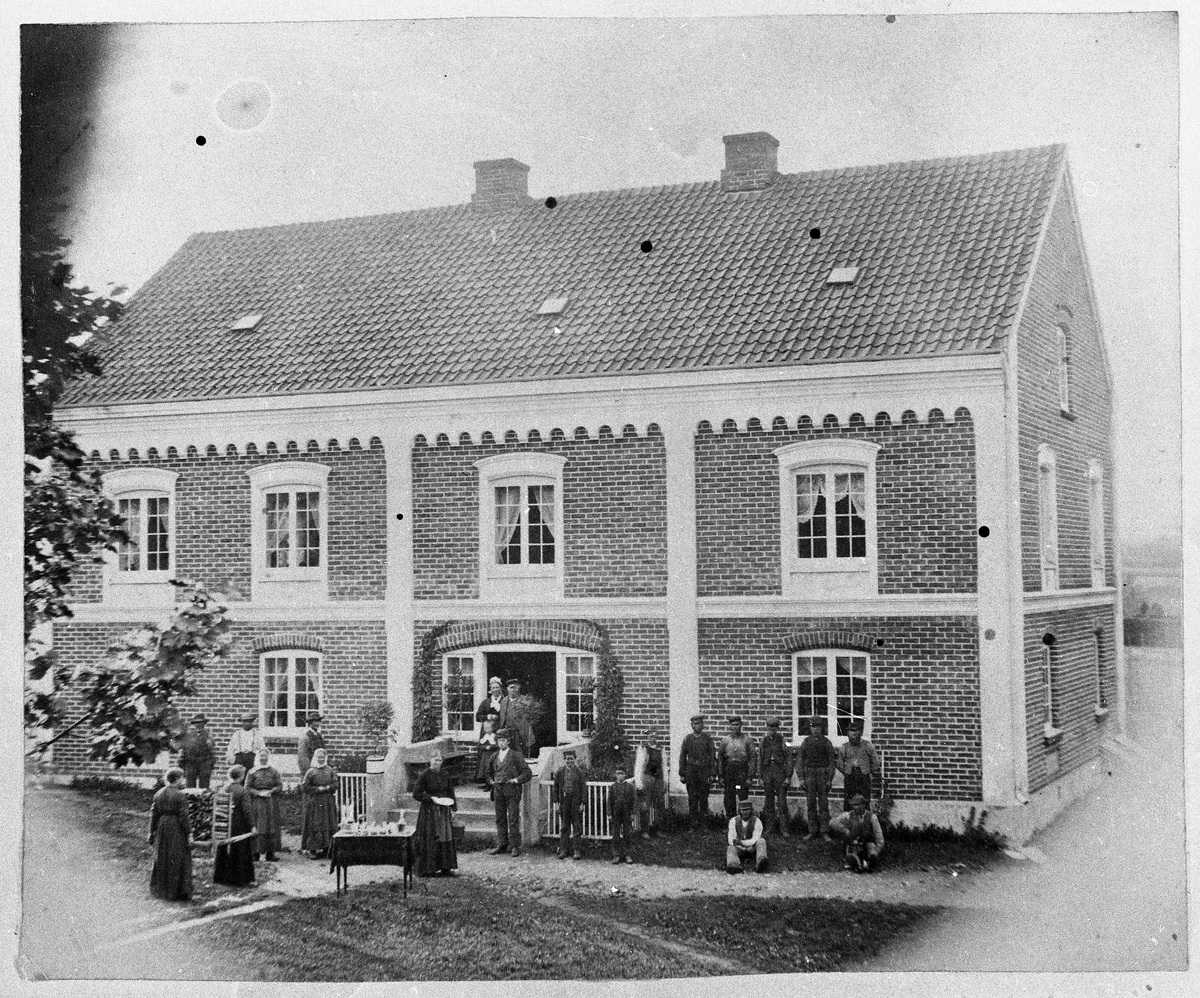 Gruppebilde tatt foran hovedbygningen på Øvre (Søndre) Hammerstad i Totenvika ca. 1878. I døra står Marie og Ole Jensen Hammerstad, trolig med barnebarnet Marie Evensdtr Rognerud (født Gihle, 1874). De øvrige personene er ikke identifisert.