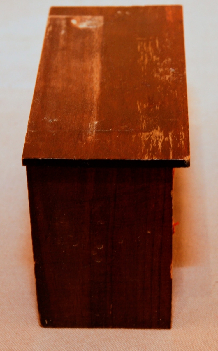 Skåp till dockskåp i mörkt trä från mitten av 1800-talet. Med guldfärgat tryck som ska föreställa fronterna av de översta två lådorna. Dörrar saknas. I tomrummet ligger 3 miniatyrböcker (FMH.05064). Det saknas ett parti/en uppsats som varit fastskruvat på ovansidan.