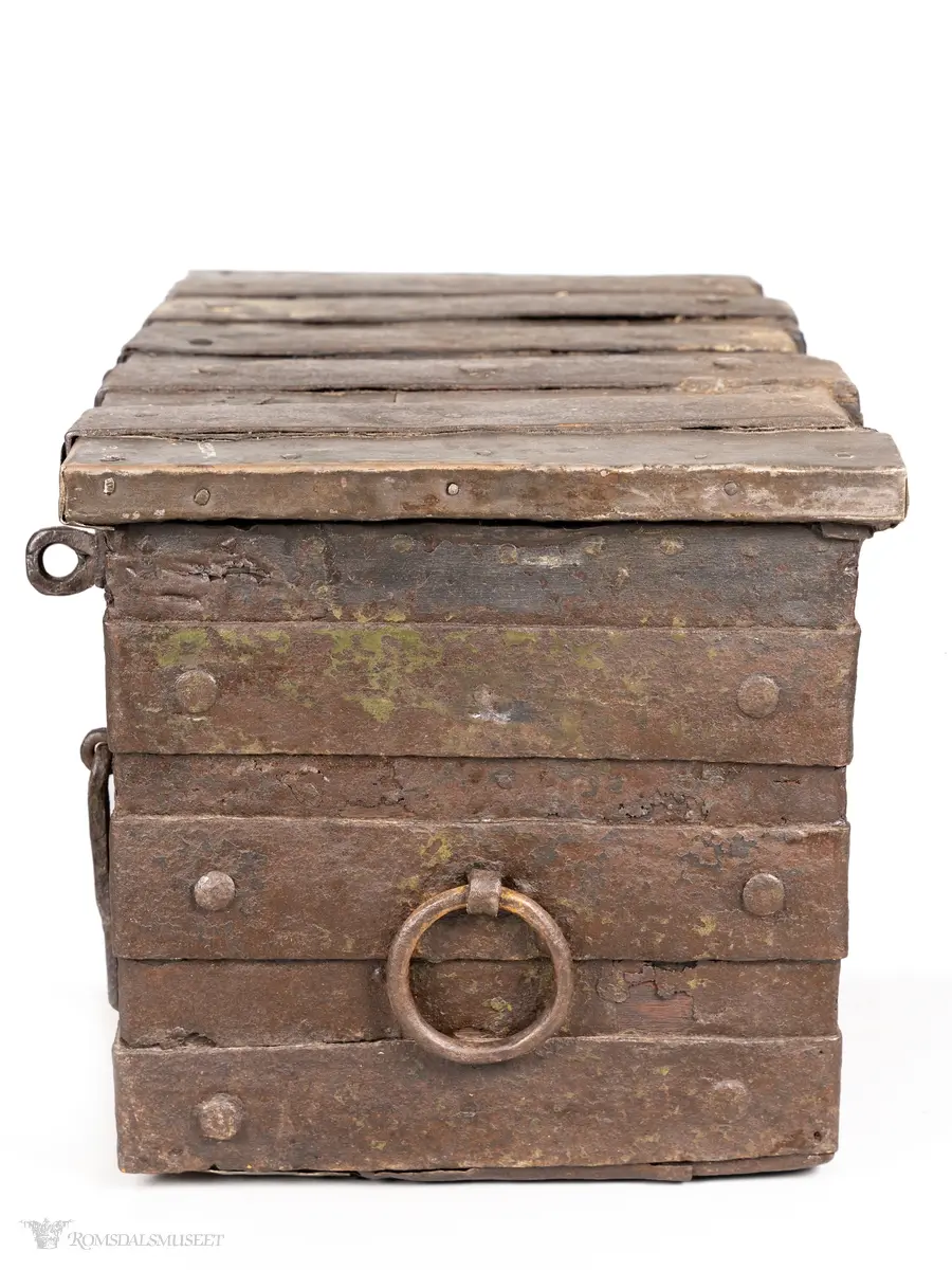 Solid kiste av tre, beslått med jernbeslag og jernplater. På høyre side er en jern- ring som benyttes som en slags nøkkel i tillegg til en annen nøkkel.