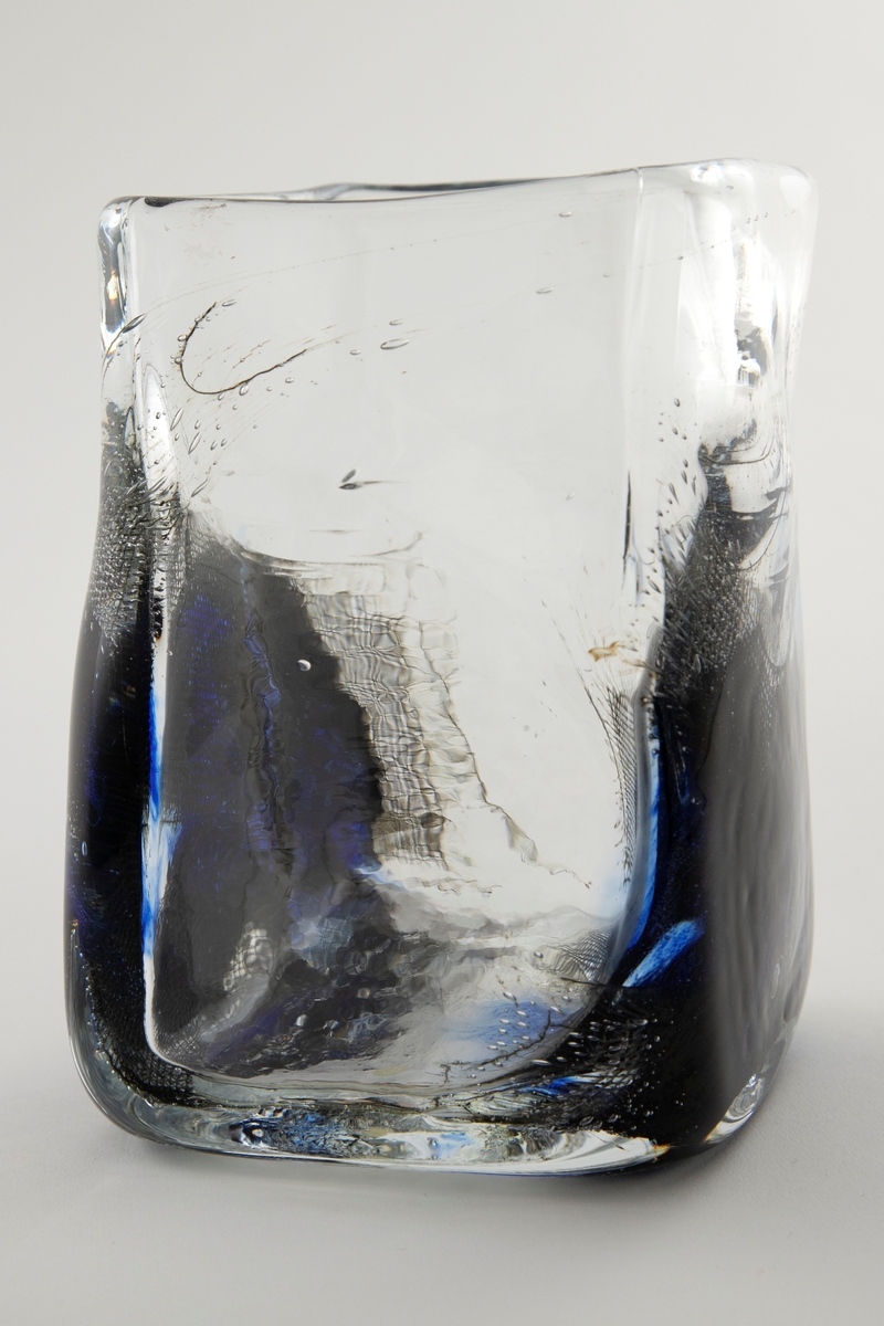 Kvadratisk vase i klart glass med tykk bunn. Nedre del er delvis utbulet, og dekorert med metallnett, luftbobler og koboltblå partier. Øvre del er utført i klart glass med enkelte luftbobler.