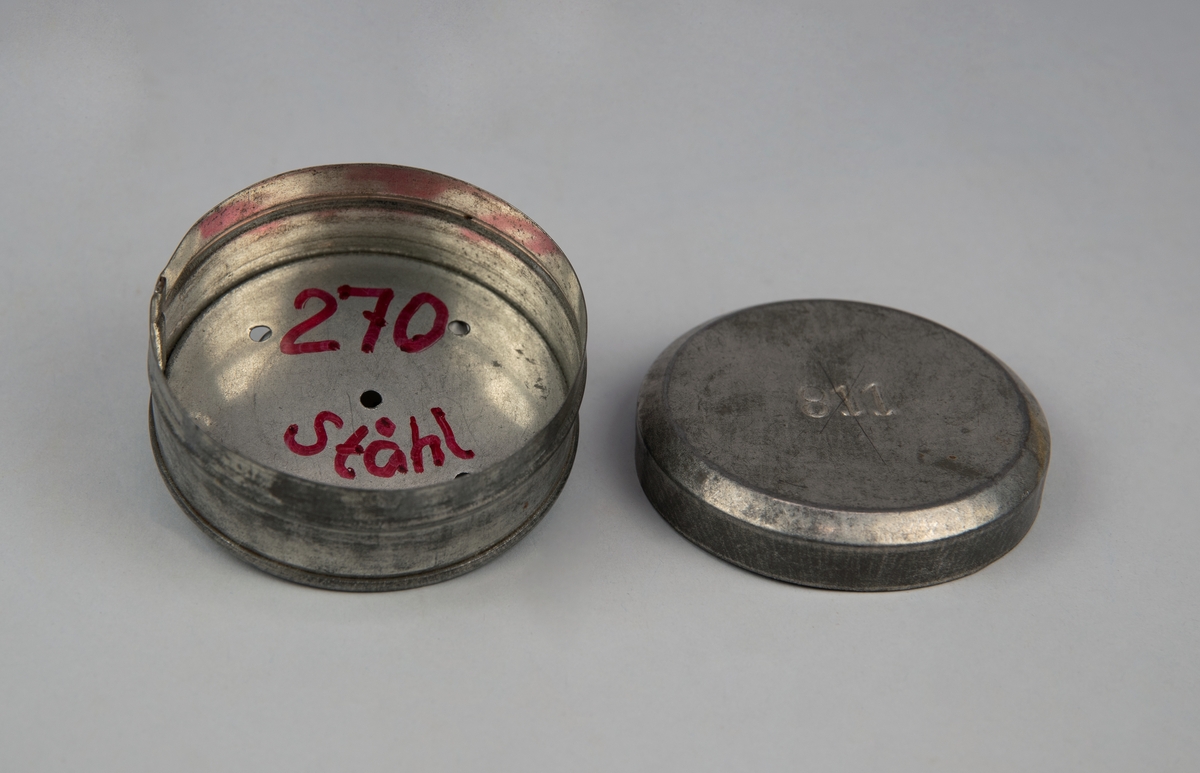 Rund avlöningsask (:1) med lock (:2) av plåt. Locket märkt "811". Asken har fem hål i botten. Insidan av asken märkt "270 Ståhl" med röd penna.