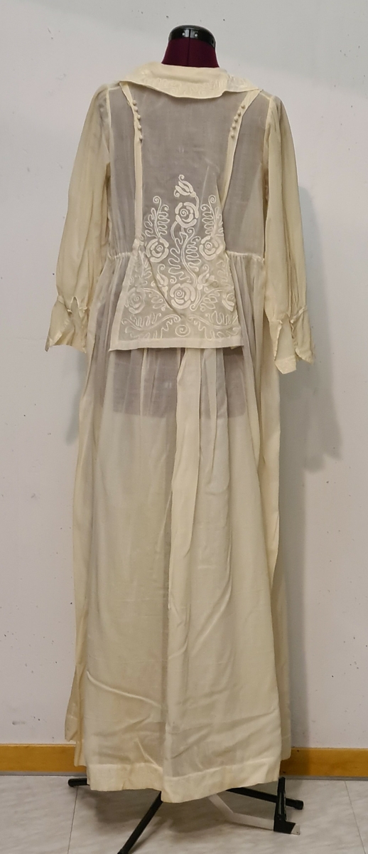 V-ringad klänning med brodyr i vitt.  Virkade runda knappar, liten broderad krage bak. Sydd till bröllop 1911. Från Sörmland.