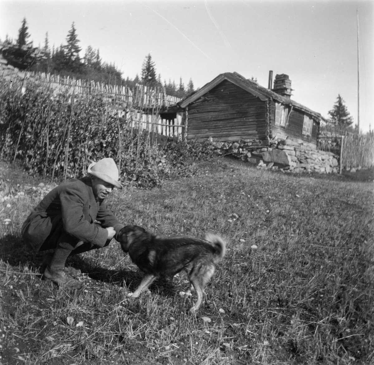 En mann og en hund foran stugbygningen i Høgbrenna i Flatmogredi i Lom. I bakgrunnen gjerder og granskog