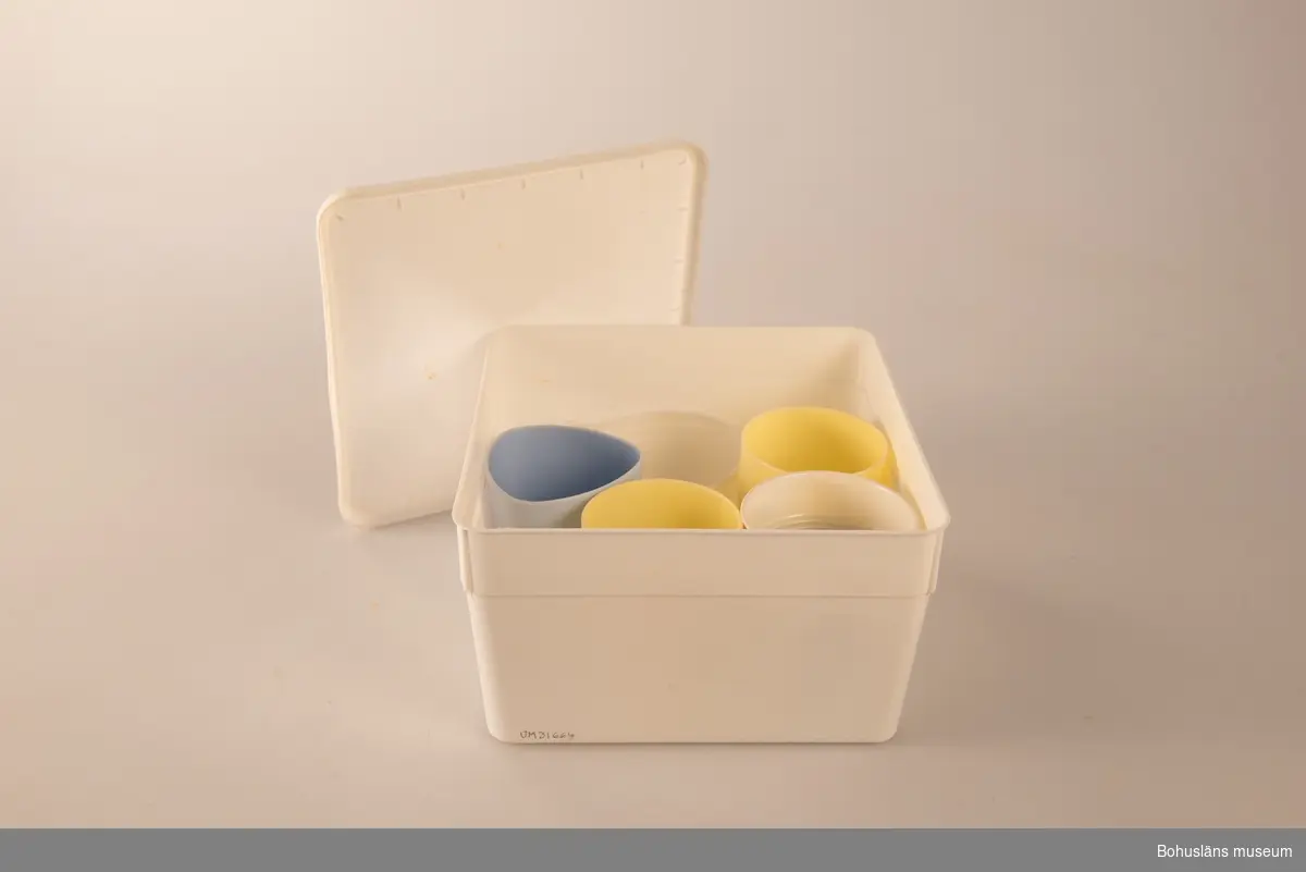 Kvadratisk låda av vit plast med plastlock. I lådan förvaras sex muggar/koppar: två gula plastmuggar, en ljusblå plastmugg, en vit plastmugg med handtag,  en engångsmugg av plast och en engångsmugg med handtag av plast.