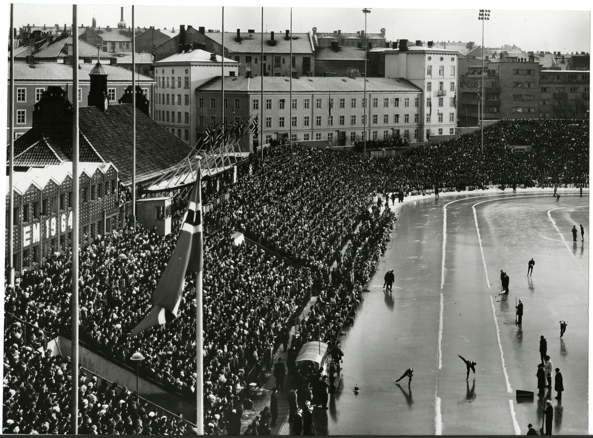 Publikum på storestå på Bislett stadion under EM på skøyter i 1964. To skøyteløpere konkurrerer på skøytebanen.   "EM på skøyter 1964- Lengdeløp var en stor folkeidrett i Norge. Bislett var hovedarenaen og samlet mange entusiastiske tilskuere ved de internasjonale mesterskapene. Det ble arrangert ett OL, 13 VM og 10 EM her før skøyteisen ble lagt for siste gang sesongen 1987/88.  Europamesterskapet i 1964 ble avholdt 18. og 19. januar med 31 utøvere fra 11 land. Sovjetiske Ants Antson vant sammenlagt foran Juri Jumasjev, Sovjet og Per Ivar Moe, Norge".  -utstillingstekst "Gjennom Linsen" 