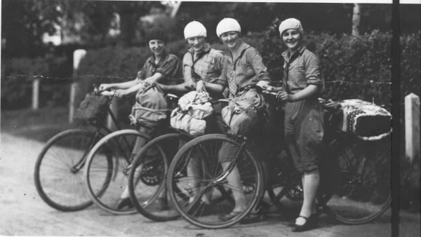 Fyra kvinnor på cykelutflykt med packning både på styre och pakethållare. Alla är iklädda knäbyxor, skjorta, slips och basker.