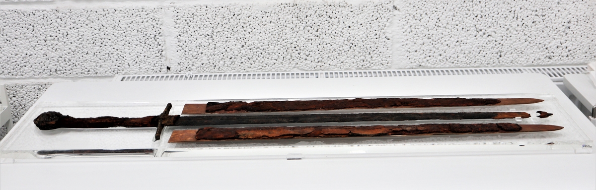 Svärd av järn med rester av trä- och läderskida kvarsittande på bladet vid upphittandet. Mycket skör. Bladet har en synlig blodränna. Rak parerstång och ett grepp som avslutas med en rund knapp som är tillplattad på ovan- och undersidan. Även på greppet finns läderrester. Svärdets metalldelar förefaller välbevarade. I samband med konserveringen lades trä- och läderskidan i två delar på var sida om svärdet.