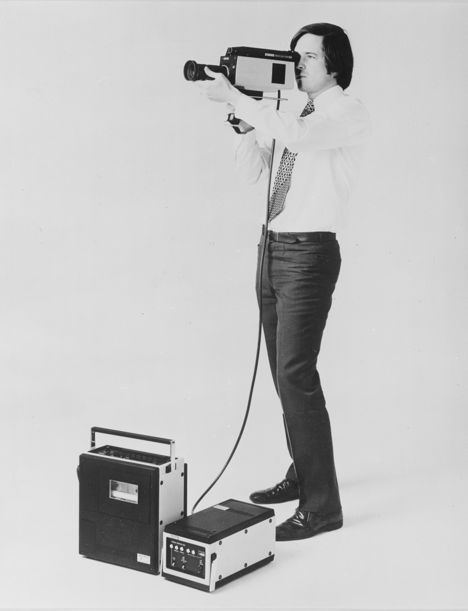 En mann filmer med videokamera. Foran ham står opptaksutstyr. På originalemballasjen står det""Videoutstyr. Acec A/S. Repro AH."