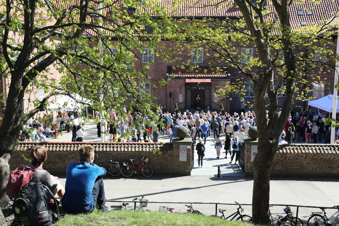 Et oversiktsbilde over Haugar kunstmuseum og borggården. Inne i gården er det mange mennesker. Det er sol og sommer. Helt i forkant på bildet, til venstre, sitter to personer med ryggen til og ser mot bygget til kunstmuseet. De sitter på gresset på en forhøyning