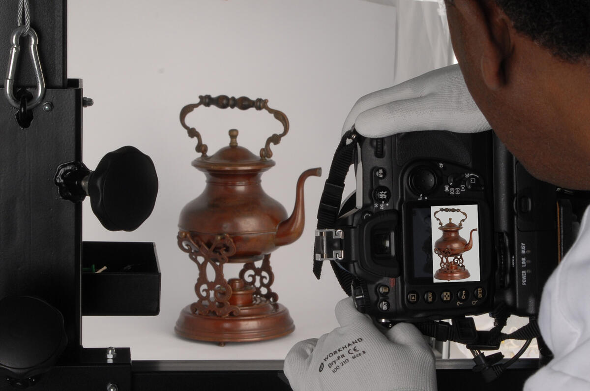 Vi ser bakhodet og en hanskekledd hånd til en mann som fotograferer en gammel kaffekanne på stativ.