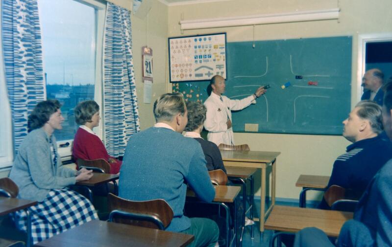 Bildet viser en lærer i hvit frakk som står og peker på en grønn tavle, Elevene ser ut til å være unge voksne.