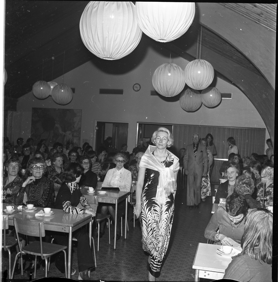En kvinnlig mannekäng skrider fram i en gång mellan bord med publik och kaffekoppar. I taket hänger flera lampor i form av stora runda bollar.