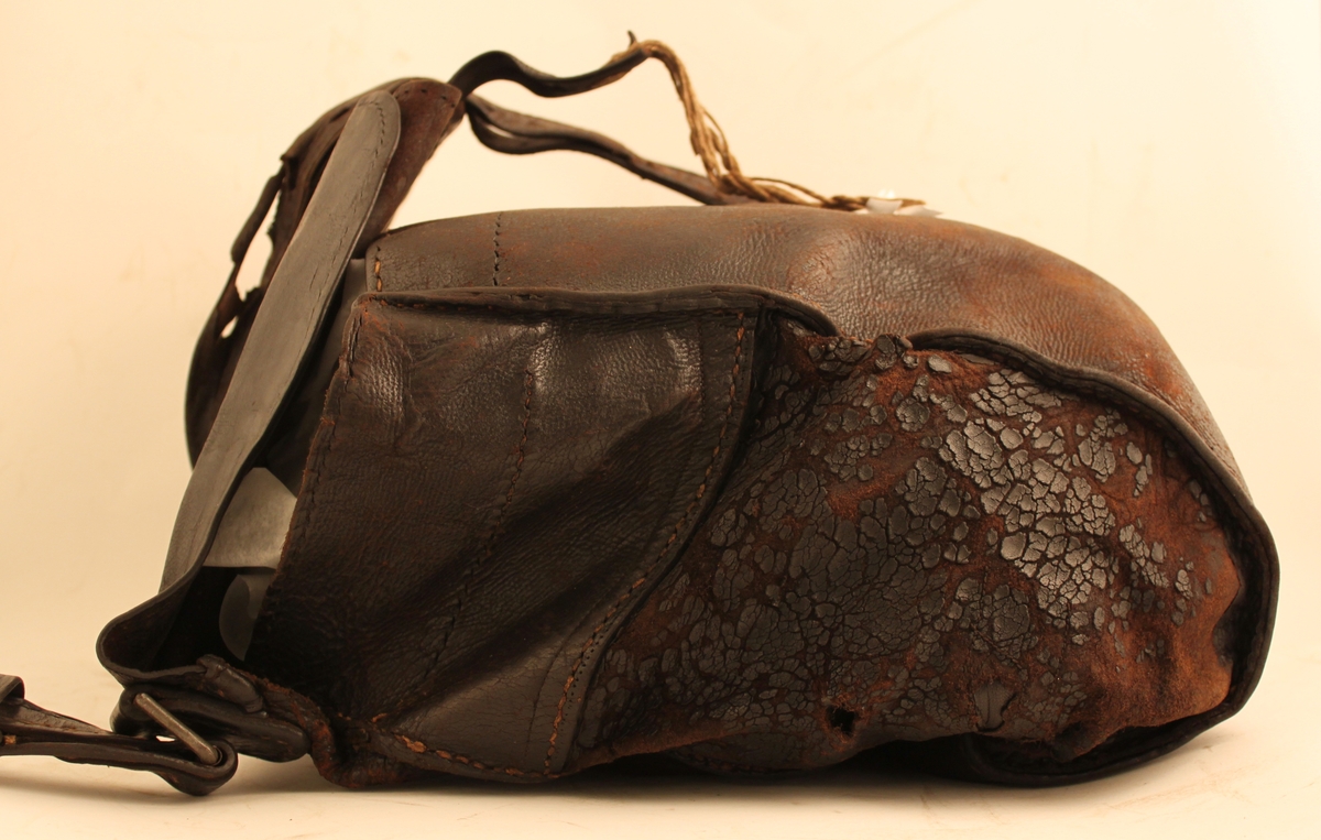 Brevbärarväska av svärtat läder med bärrem och spänne. Inuti väskan på baksidan två mindre fack. 

Ett knippe snören fastbunda vid spännet. Hjälp för att stänga väskan?

Lädret slitet.