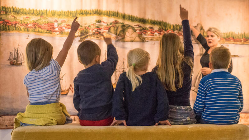 Bildet viser ryggene til barn som sitter på en benk. Mange av dem har en hånd i været. De ser på et stort maleri og en kvinne som står og snakker. (Foto/Photo)