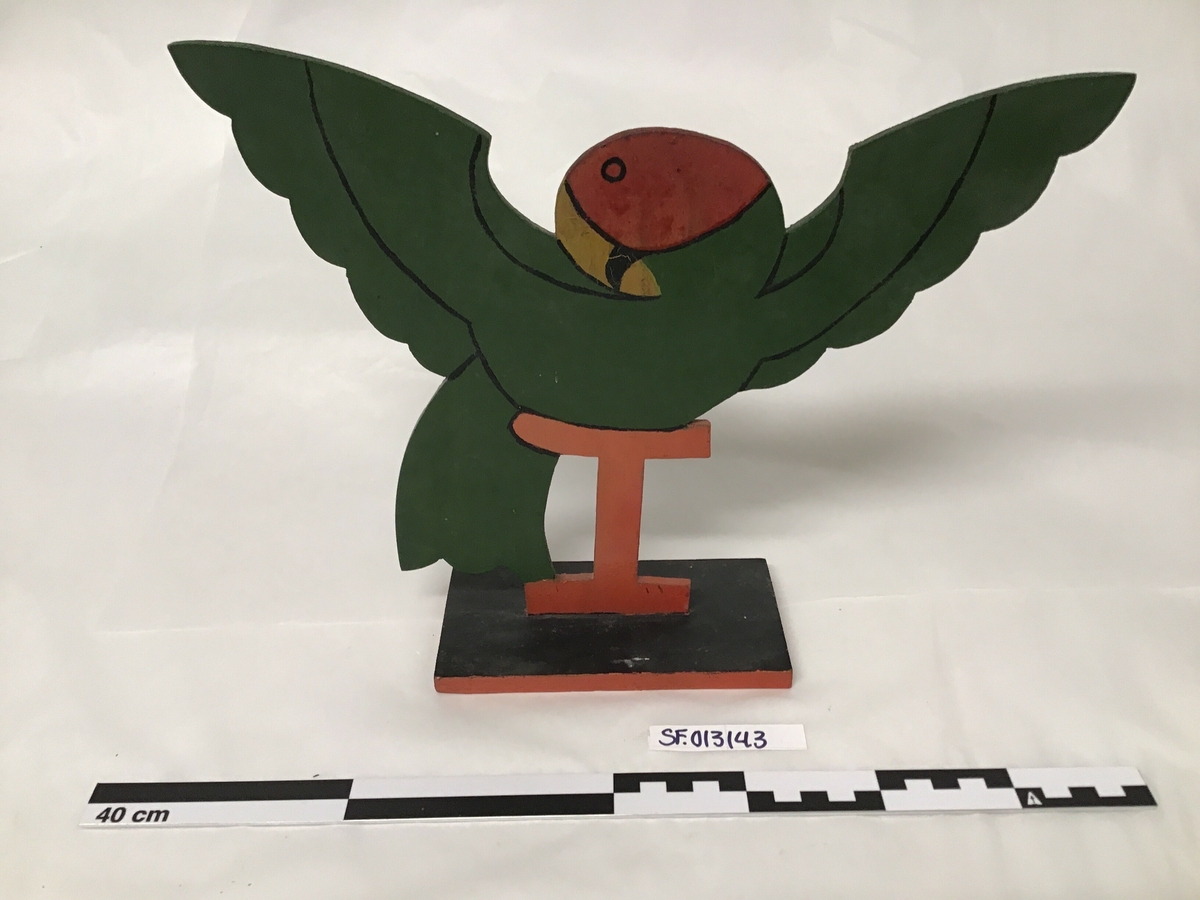 Utskoren trefigur, forma som ein papegøye som sit på ei vagl.  Laga av ei treplate og festa på ei fjøl som dermed fungerer som ein sokkel.