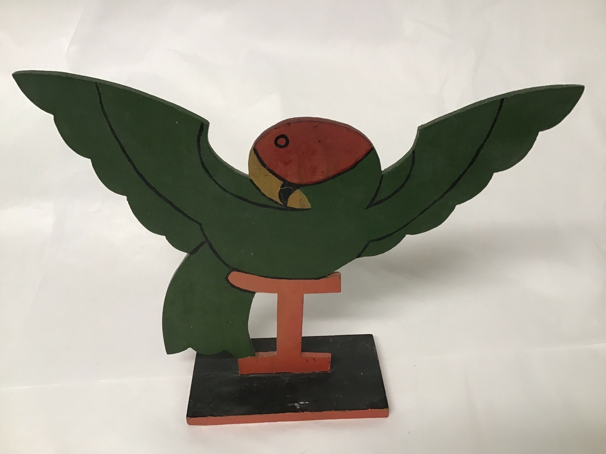 Utskoren trefigur, forma som ein papegøye som sit på ei vagl.  Laga av ei treplate og festa på ei fjøl som dermed fungerer som ein sokkel.