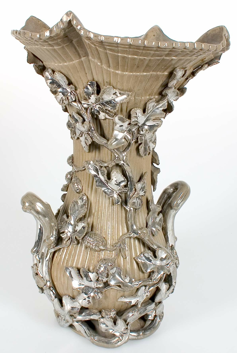 Vas av gråglaserad keramik med ekollon och ekblad i relief överdragna med silver eller kromglasyr. Innevas av ofärgat glas, strutformad med en ögla nedtill, längd 22 cm, diam 14,5 cm.