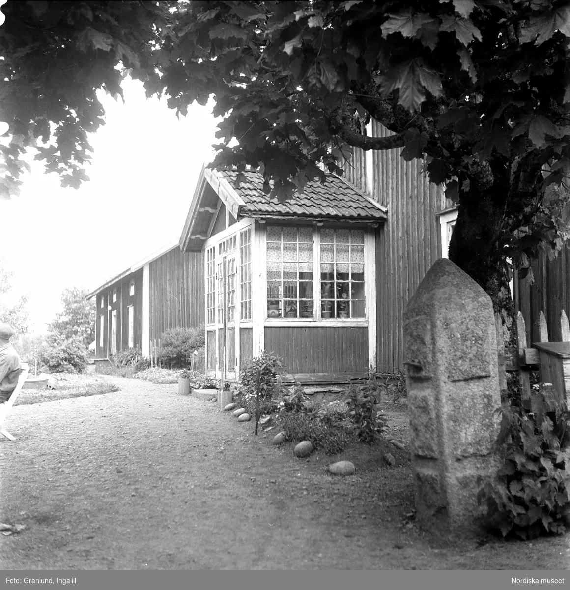 Bostadshus i byraden i Kullarp, Ydre härad, Östergötland. I förgrunden en gridstolpe av sten och ett träd. I vänsterkant anas en man sittande på en trädgårdsstol.