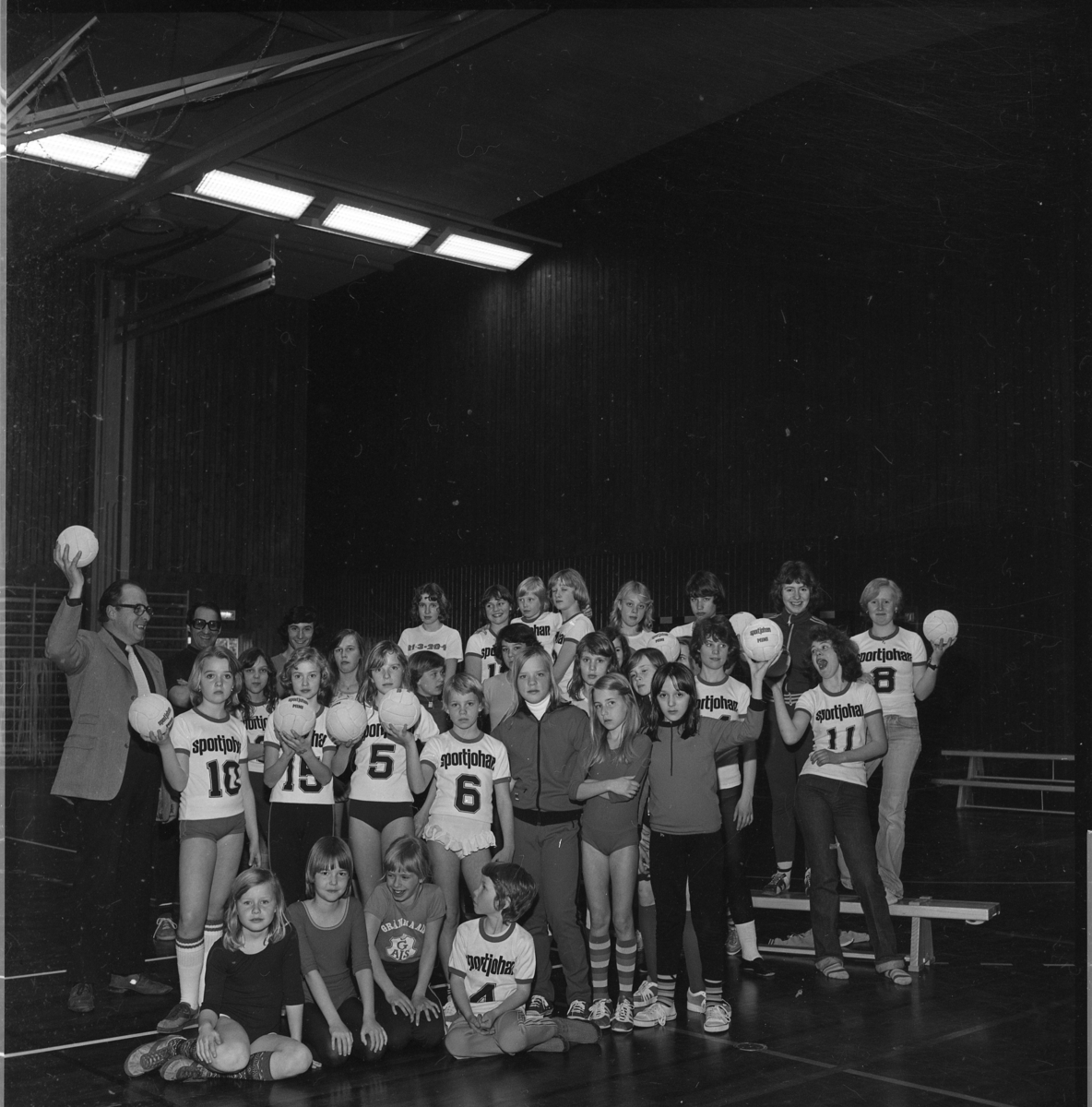 Ungdomar i Gränna AIS handbollssektion står uppställda med handbollar i händerna i Ribbahallen. De bär t-shirts märkta "Sportjohan" och en siffra. Två män står till vänster. Det är representant för Rotary Gränna respektive handbollstränaren.