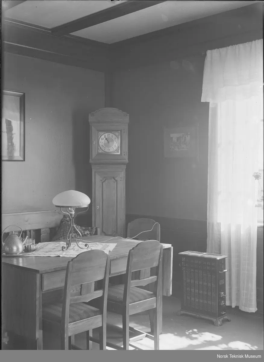 Spisebord med elektrisk lampe, gulvklokke og elektrisk varmeovn i bakgrunnen; interiør fra mønsterbruket på NEBB's jubileumsutstilling i 1914