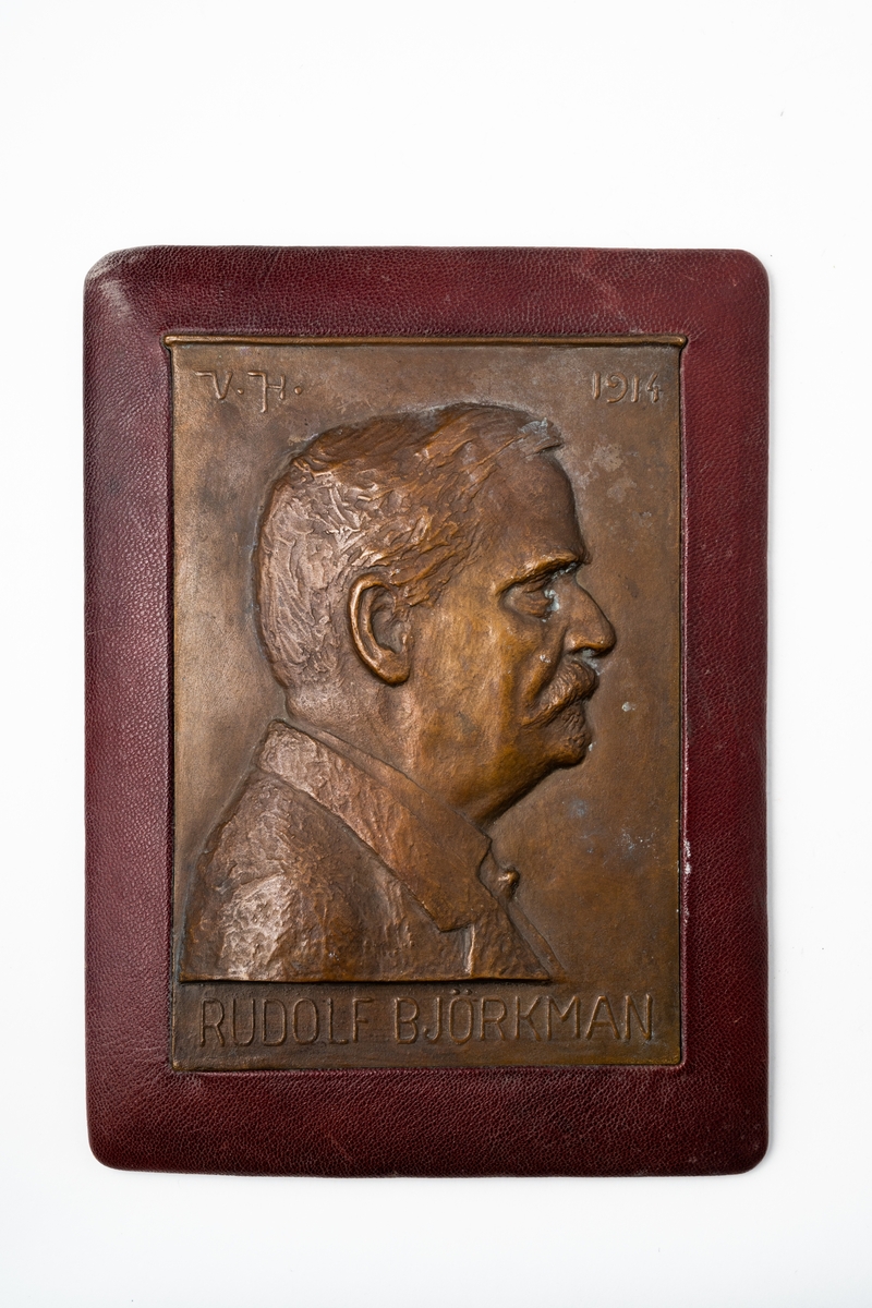 Plakett i brons, monterad på träplatta klädd med tunnt skinn, med bild av redaktör Rudolf Björkman i profil. På baksidan är ett maskinskrivet papper fastsatt med text om Rudolf Björkman.