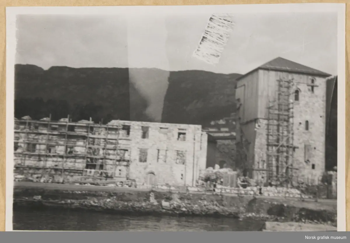 Bygninger med stillaser rundt. Bergenhus Festning, trolig restaurering etter eksplosjonsulykken på Vågen 20. april 1944, som medførte svært omfattende skader på festningsbygningene. Fotografert i forbindelse med Vestlandsk Trykkerstevne i Bergen, 1946.