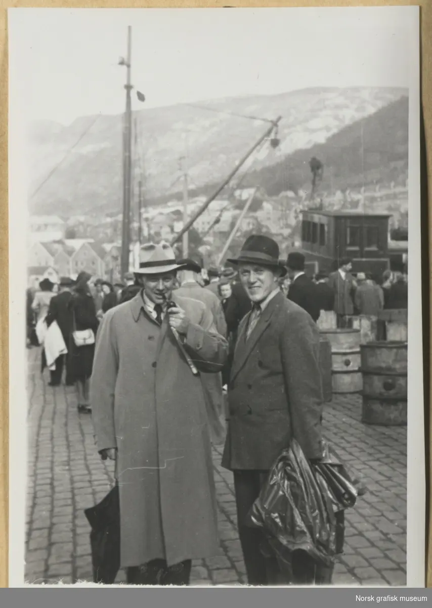 Havnebilde. To menn i dress, frakk og hatt har stilt seg opp foran kamera. I bakgrunnen ser vi mange mennesker, båter / båtmaster og tønner, og landskap med bebyggelse. Fotografert i forbindelse med Vestlandsk Trykkerstevne i Bergen, 1946.
