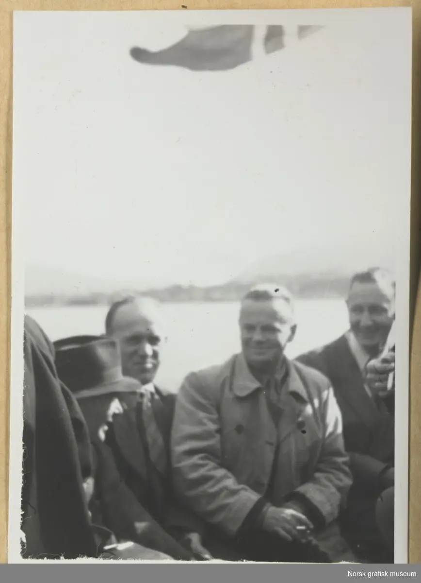 Gruppebilde, utendørs. Fire menn, tre av dem med dress og slips, den fjerde ser vi bare hodet av, med hatt på. I øverste bildekant skimtes et vaiende norsk flagg. I bakgrunnen kystlandskap med åser. Fotografert i forbindelse med Vestlandsk Trykkerstevne i Bergen, 1946.
