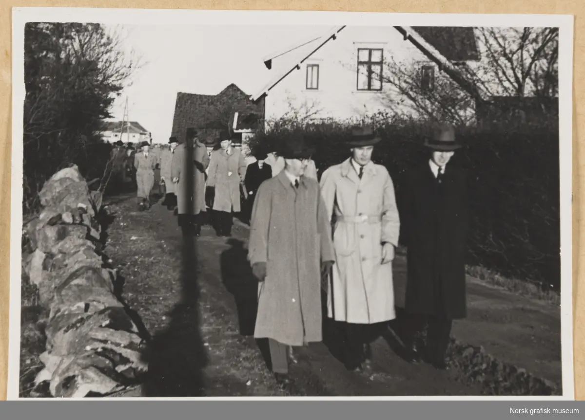 Utendørsmotiv. En gruppe menn i hatt og frakk kommer gående mot kamera langs en vei. På venstre side er det en steinmur, på høyre side en hekk. I bakgrunnen bolighus. Fotografert i forbindelse med Vestlandsk Trykkerstevne i Stavanger, 1948.