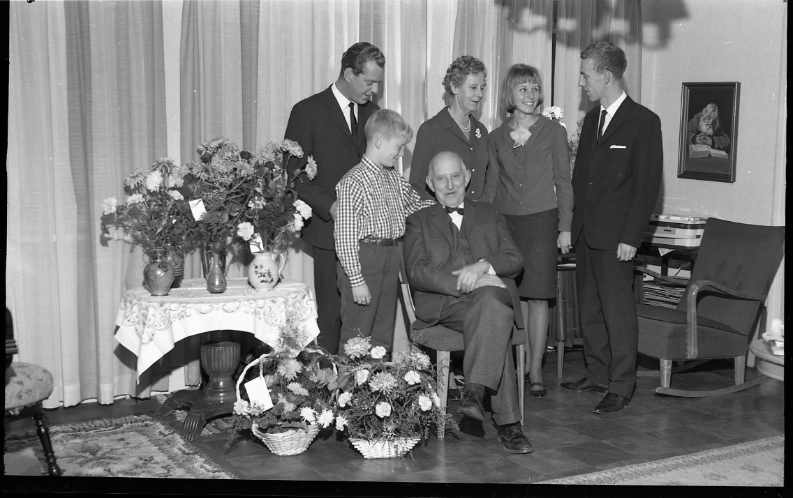 En äldre man sitter i en stol omgiven av barn och barnbarn. På ett bord intill och på golvet står blomsterbuketter. Den äldre mannen heter Möller och fyller 75 år. Männen till vänster och höger är sönerna Stig respektive Rolf.