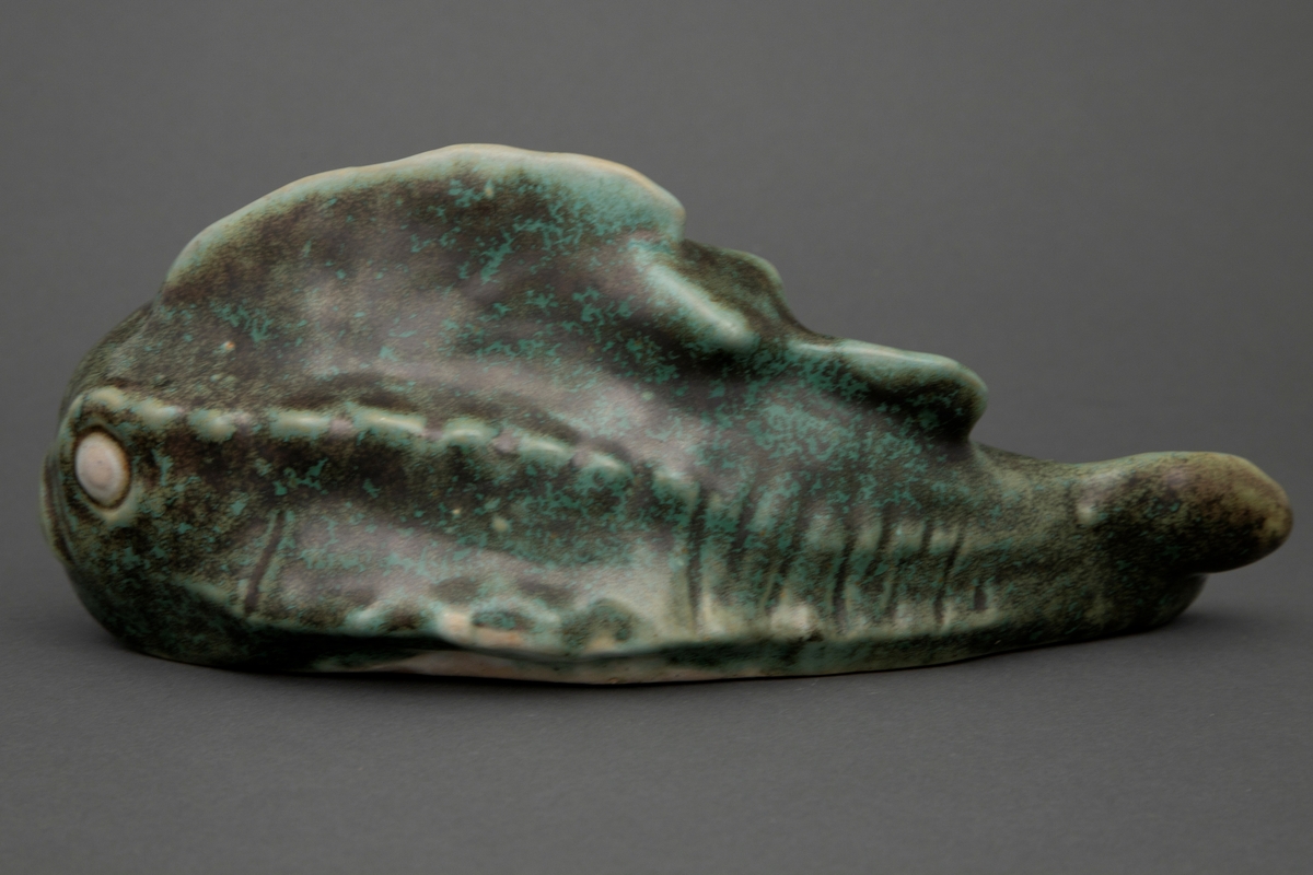 Figur som viser en fisk. Figuren i støpt steingods med grønn kobberglasur.