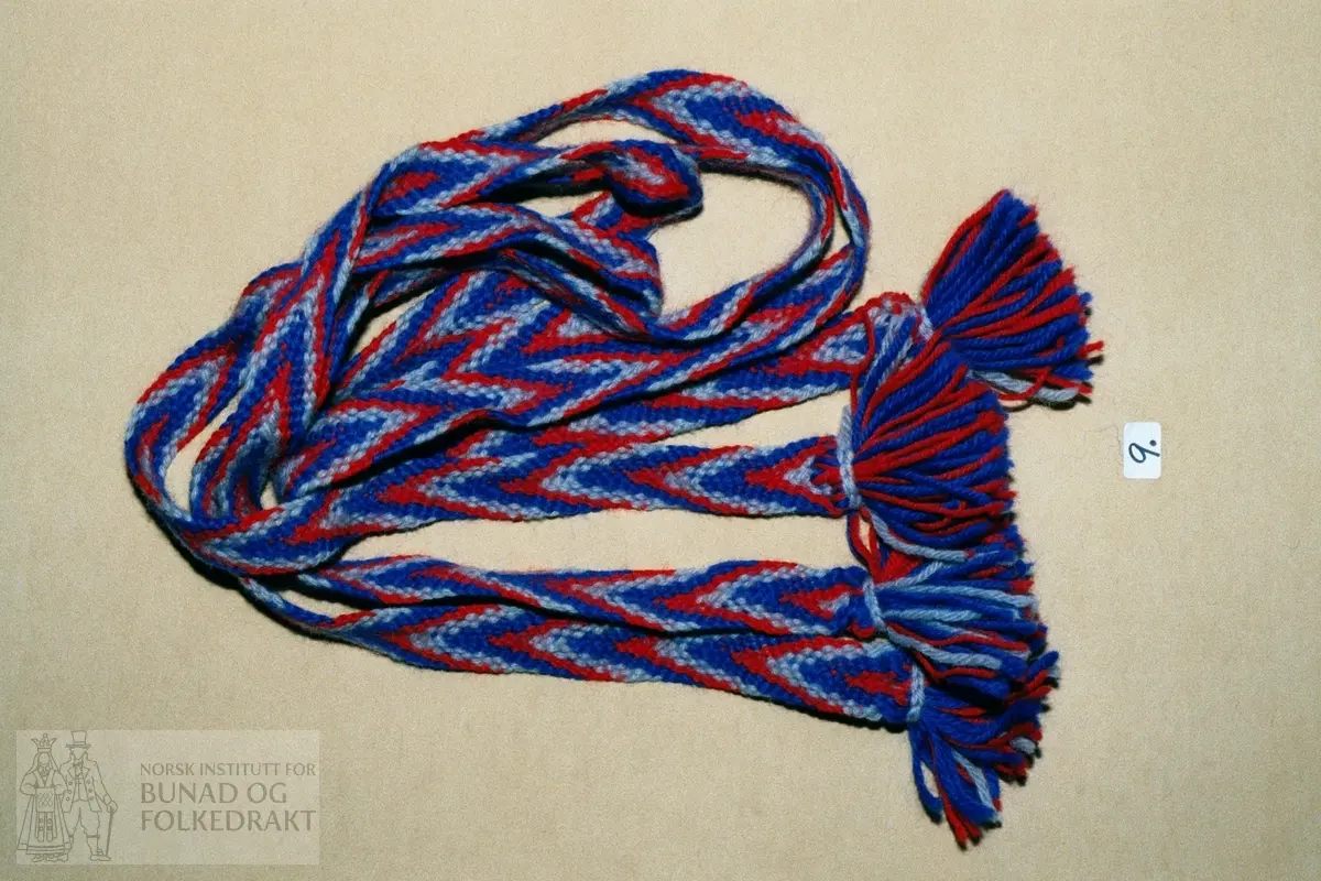 Fingra med tretråds ullgarn i raudt, blått og lyseblått.  22 trådar.  Lengde:   144 cm. Breidde:  2,4 cm.