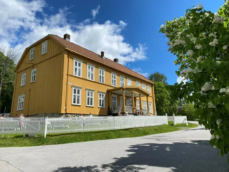 Aamodtgården i sola. Gult, staselig bygg med to etasjer og et loft. Huset og hagen er inngjerdet av et hvitt stakittgjerde. Til venstre i bildet, i forgrunnen, er det en syrin med hvite blomster. Fotografiet er tatt i fint sommervær.