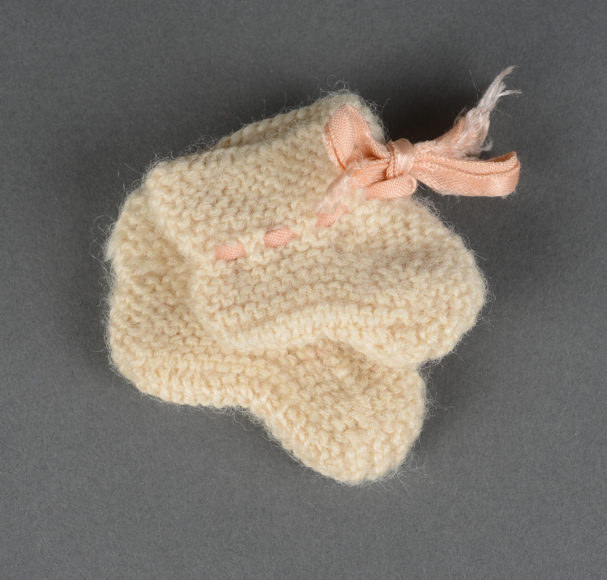 Et par strikkede dukkesokker. De er strikket i rettstrikk. Vend ankelhøyde er det strikket en hullbord, og i den ene sokken er det tredd i en rosa silkesløyfe.