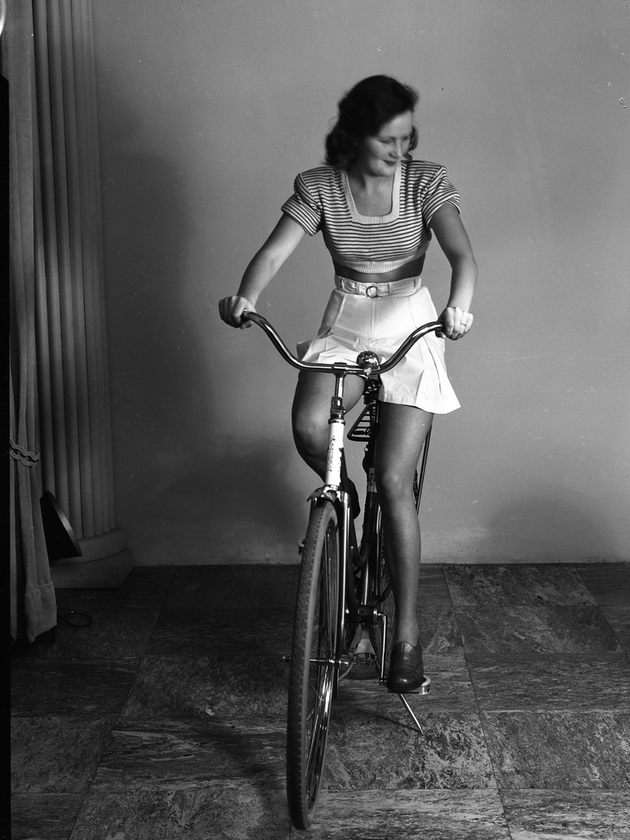 Sannolikt reklamfotografi för cykel (Monark) då skärpan ligger på styret. En kvinna sitter på cykel med stödet nedfällt i fotostudio.