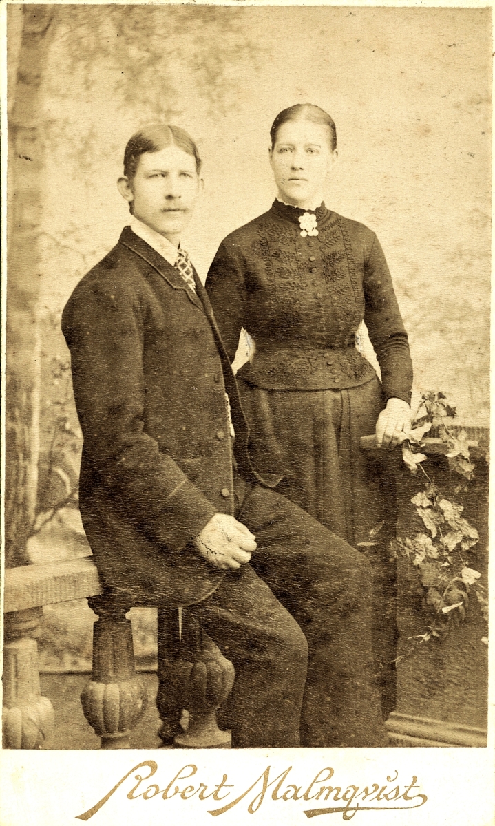 Ett ungt, okänt par i mörka kläder, troligen fotograferade i Halmstad. Mannen sitter på en balustrad och har en strikt mittbena och rutig slips. Kvinnan bär en rikt dekorerad blus med en stor brosch under hakan.