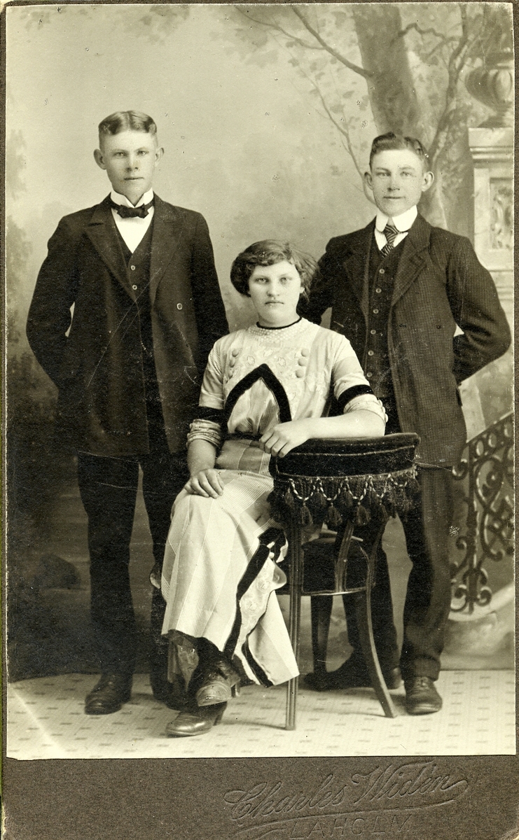 Tre okända ungdomar, sannolikt syskon, fotograferade i Laholm. Flickan sitter på en stoppad stol med fransar och de två ynglingarna stående på ömse sidor om henne. Hon bär en ljus klänning med klädda stora knappar och breda, svarta band på liv, ärmar och kjol.