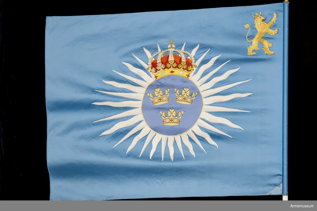 På blå duk i mitten lilla riksvapnet omgivet av vita strålar och tungor. I övre inre hörnet förbandets vapenbild; ett med öppen krona krönt gult upprest lejon med röd beväring.