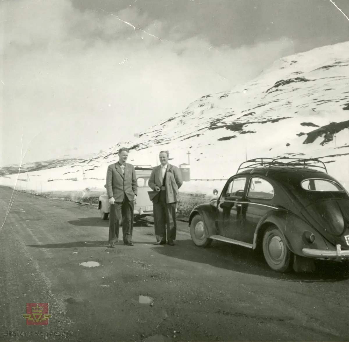 Befaring andre mai 1958. VW Boble fra 1953-1957. Regnr. E-1398. 
I bakgrunnen Statens vegvesen varebil, registreringsnr. E-16477. Bilen er en Opel Olympia 1952-1953 modell.
Bak bilene skimtes KNA Veitelefon. 
Personen fra venstre er ukjent, til høyre står oppsynsmann Kristen Eriksen.