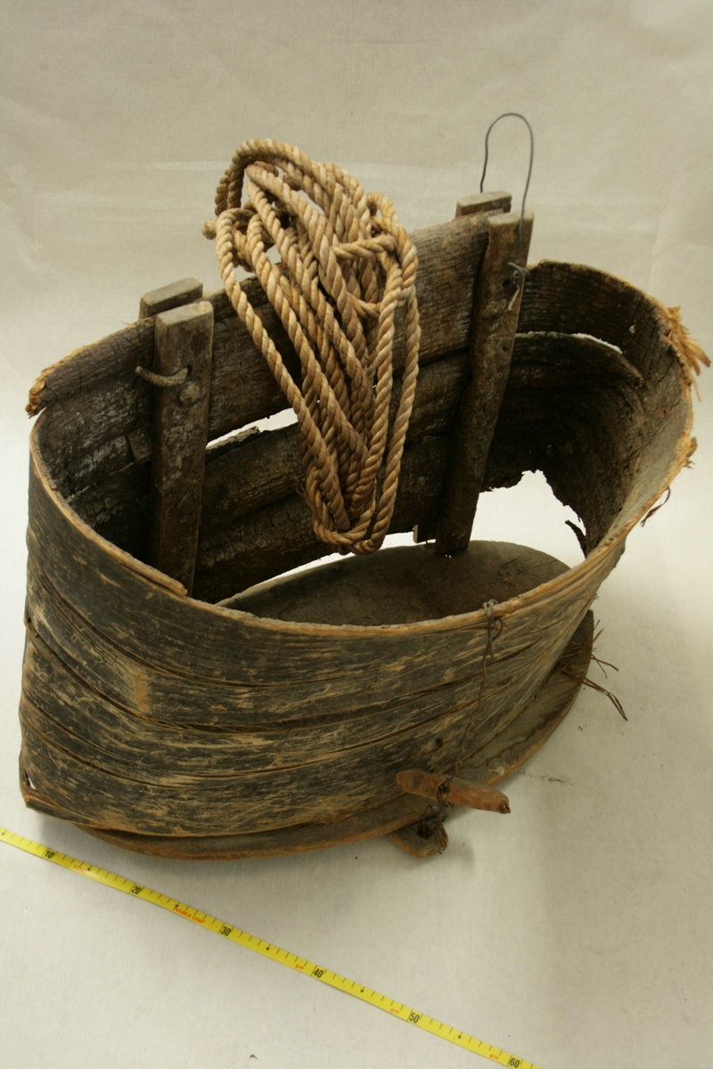 Stor laup laget av bark som er bøyd med utsiden innover, trebunn, to fester til tau, noen meter med tau ligger oppi laupen