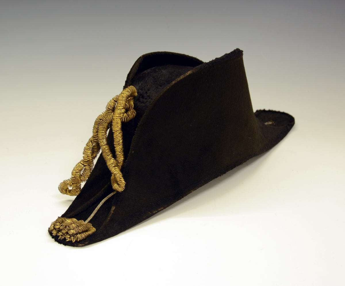 TGM-BM.1940-41:038.A Tosnutet hatt.
TGM-BM.1940-41:038.B Hatteeske
Fra protokoll:Snutehatt av filt, med vidne gullband feste til "løveknappar". Til hatten høyrer blekkøskje.

