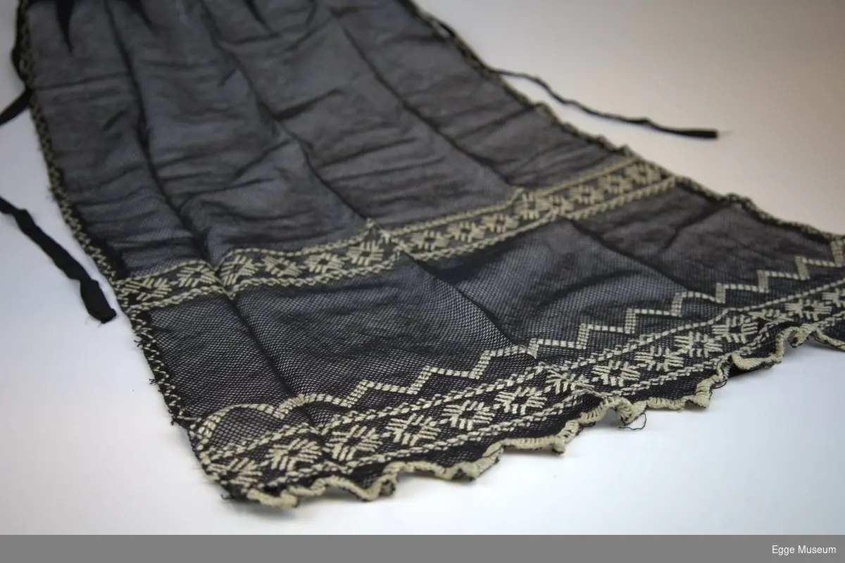 Sort forkle av tyll/bomullsnett med brodert dekor sydd med silkegarn. Knytebånd av sort silke.