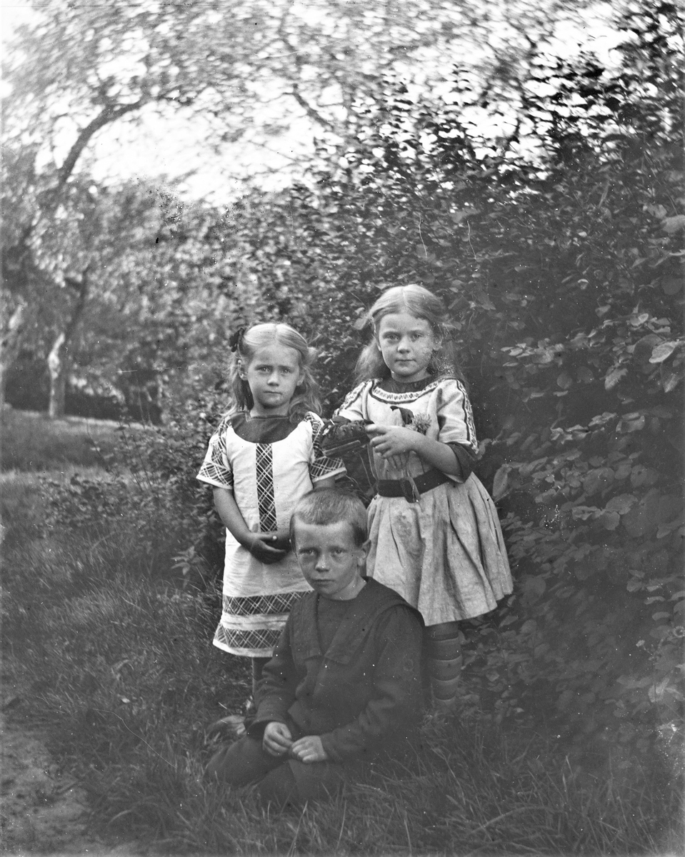 Tre små barn, troligen syskon, fotograferade i en trädgård. Flickan till höger har en påsnörad skena på ena armen. (Se även bilderna GEB180, GEB183)