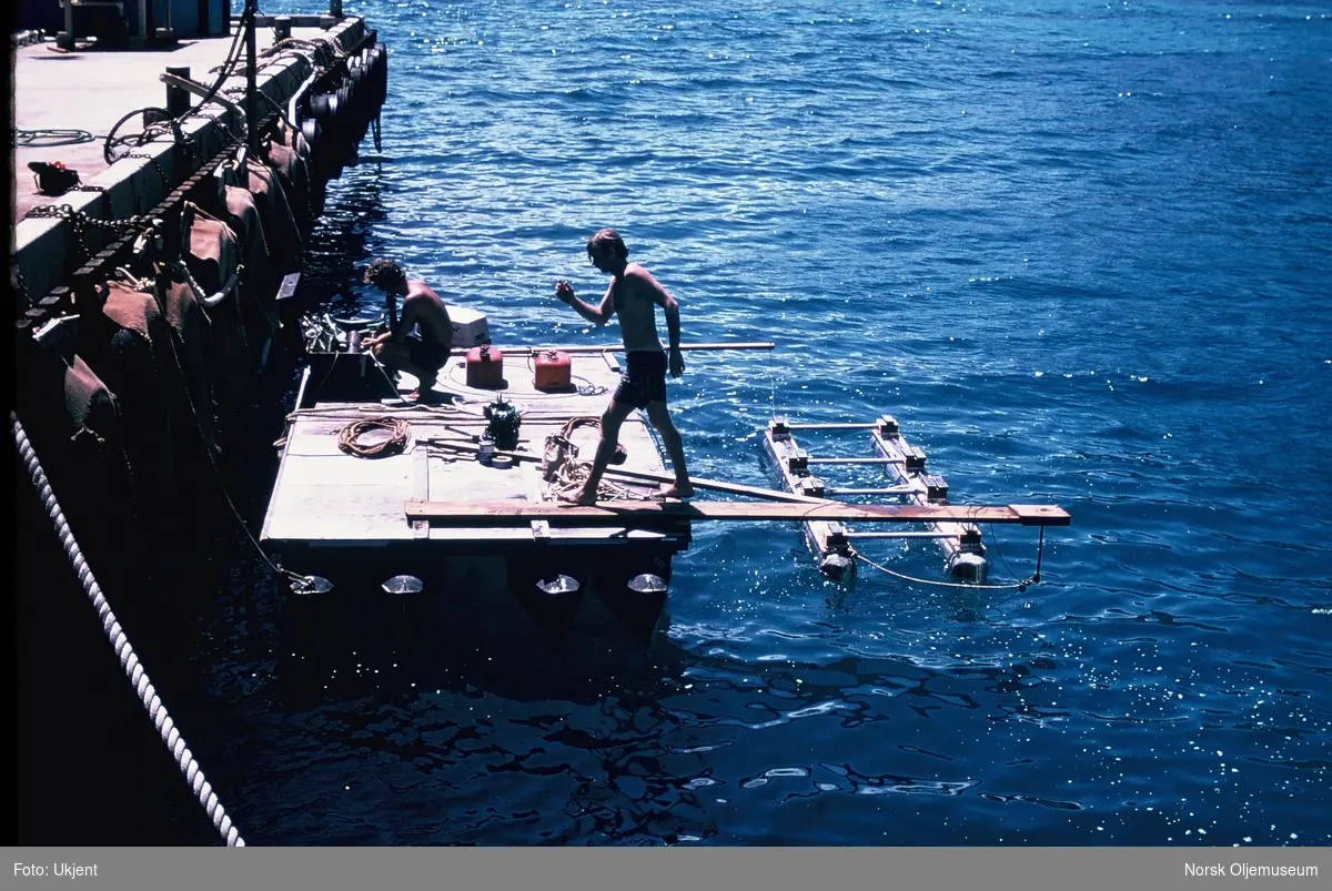 Test av Shell sin "SPRS" i sjøen. Farkosten er fortøyd til planker. To menn jobber med testingen fra ei flåte. Han ene har vært i vannet.