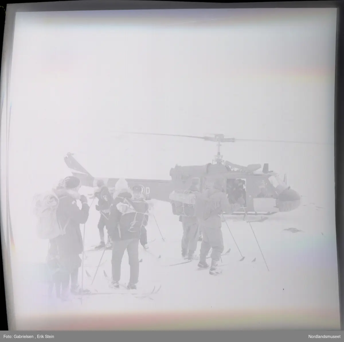 Fotografi av mange menn og kvinner kledd i vinterklær
med sekker på ryggene som står på en snøslette med ski på seg i næreheten av et helikopter som man
ser en mann inni i døråpningen til helikopteret 
som man ser står på snøsletten ved et berg