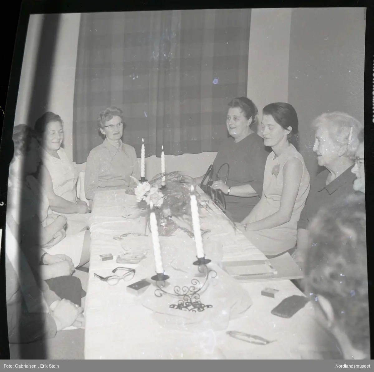 Fotografi av noen kvinner som sitter på stoler rundt
et bord som det står noen stearinlys på og en plante 
med blomster i en vase på bordet og noen fyrstikkesker som
ligger på bordet og briller og en papirbunke og et en bok
og et vindu med gardiner med mønster som er trukket for
i rommet og man ser at kvinnen som sitter lengste framme
til høyre ved bord holder en svart veske i fanget sitt