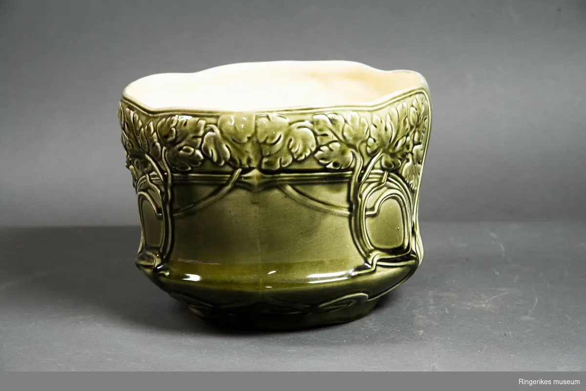 Stor potteskjuler i keramikk. Dekorert med ranker med blad som minner om vindrueblader. Glasert. Olivengrønn på utsiden, svært lys gul på innsiden.